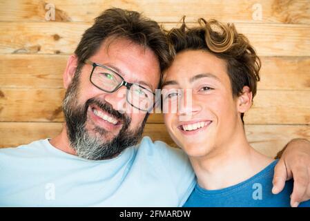Head shot happy teenager son embracing shoulders of happy middle aged father, looking at camera. Positive two male generations family supporting each other, visualizing planning future together. Stock Photo