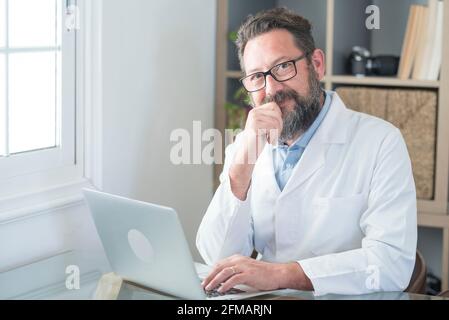 Smiling old male doctor in glasses and white uniform sit at desk in hospital work on laptop write in journal, happy mature senior man physician fill patient medical history anamnesis on computer Stock Photo