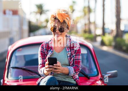 Trendy female young adult caucasian woman use modern phone connection outdoor near her pretty red car in travel and independent lifestyle - concept of app messaging and texting chat people Stock Photo