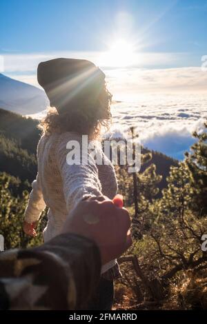 Portrait of happy young adult woman holding hand of her boyfriend while walking by mountain stream. Couple enjoying leisure in nature. Amazing mountain sunset in background Stock Photo