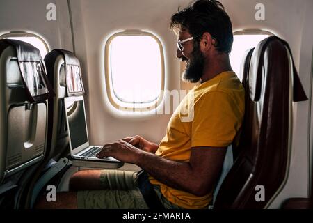 Hipster man with beard work on laptop computer sitting on board on airplane flight during travel - concept of free office remote worker and smart working lifestyle - people use connection technology inside fly aircraft Stock Photo