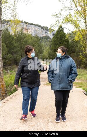 Selective focus of two middle-aged women wearing masks for Covid19 virus walking along a rural path in the countryside with trees in a sunny sunset an Stock Photo