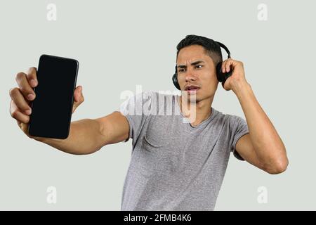 28 year old Brazilian with one hand on the wireless headphone, looking at his smartphone and showing it to the camera. Stock Photo