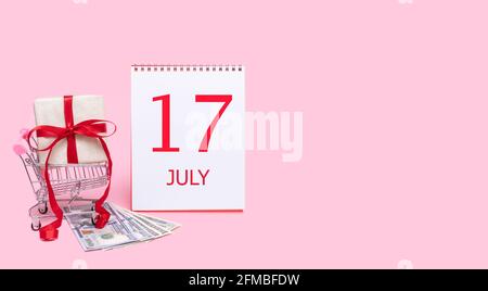 17th day of july. A gift box in a shopping trolley, dollars and a calendar with the date of 17 july on a pink background. Summer month, day of the yea Stock Photo