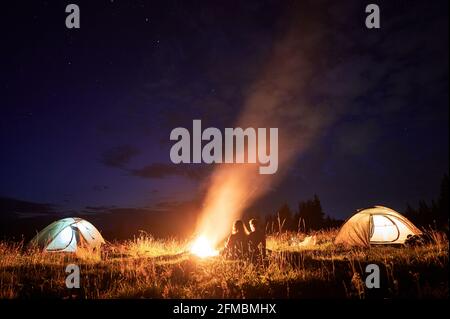 Horizontal snapshot of couple of tourists spending time together in camping. Young man and woman, sitting close near campfire in the evening outdoors in nature. Glowing tents on the sides Stock Photo