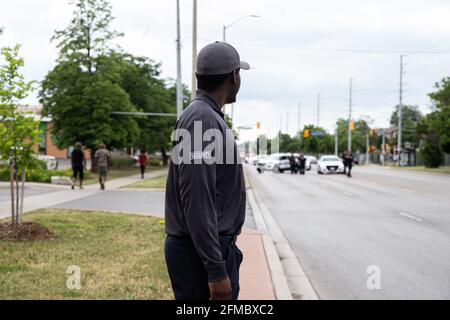 Black security guard in uniform patrolling a wide street in the daytime with police cars in the background. Stock Photo