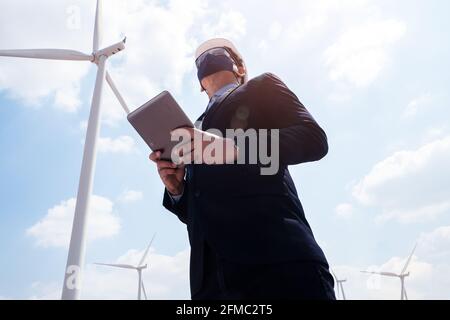 Successful engineer windmills manager wearing face mask  and using tablet with wind turbine in background Stock Photo