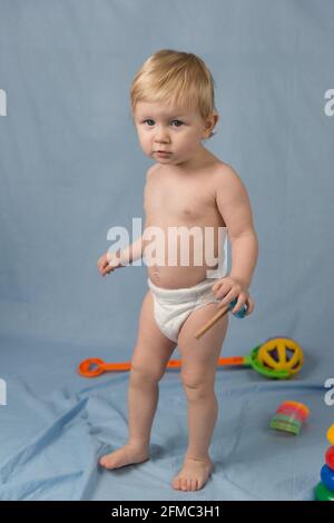 A little girl 1 year old in a white diaper walks on a blue background with colorful toys Stock Photo