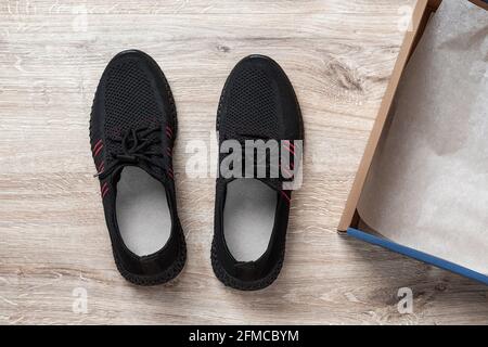Black mesh sneakers near brown cardboard box on the floor. Open box with new modern men shoes with laces for active lifestyle, fitness and sport. Stock Photo
