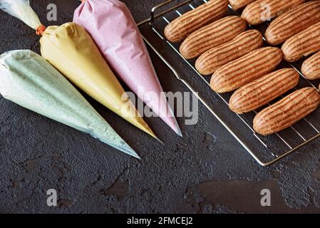French pastry dessert eclair. Freshly baked aromatic custard cakes cool on the wire rack. Pastry bags with mint, mango, berry creams. Soft selective f Stock Photo