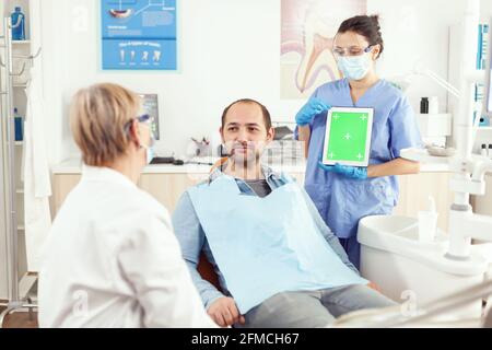 Stomatology nurse pointing her hand on mock up green screen chroma key tablet with isolated display. Sick man patient sitting on dental chair in stomatology office waiting for toothache treatment Stock Photo