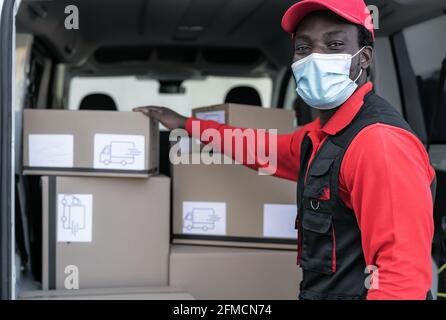 African delivery man loading boxes in van truck while wearing face mask to avoid corona virus spread - People working with fast deliver Stock Photo