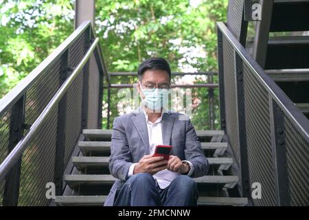 Businessman wearing face mask sitting on stairs and surfing his phone. Urban with green setting. Stock Photo