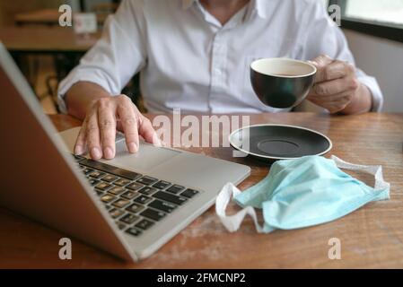 Businessman touching mouse pad on computer laptop and holding cup of tea with another hand. Face mask on table. Stock Photo