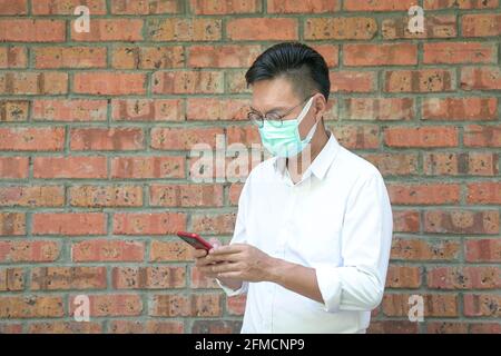 Businessman in face mask standing and using his cellphone for messaging. Brick wall background. Stock Photo