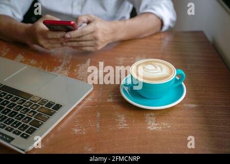 Man reading phone with computer and cup of coffee on table.  Close up view on cup of coffee in blue cup. Stock Photo