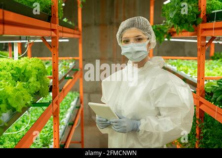 Portrait of female grower in protective suit, mask and goggles standing with digital tablet in vertical farming greenhouse Stock Photo
