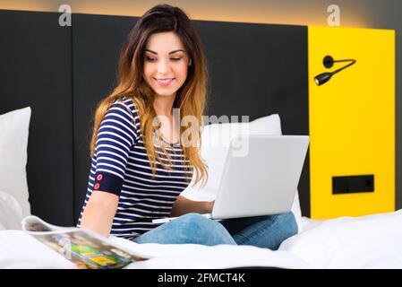 Young woman sitting on bed of a hotel room, she is on vacation and using the wifi in the room for internet with the computer Stock Photo