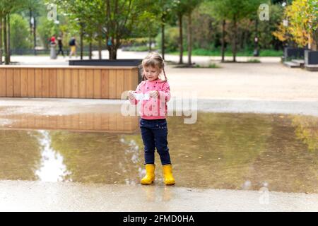 a little girl in a pink sweater and rubber boots launches a paper boat in a large puddle Stock Photo