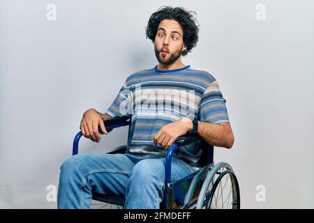 Handsome hispanic man sitting on wheelchair making fish face with lips, crazy and comical gesture. funny expression. Stock Photo