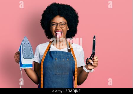 Young african american woman dressmaker designer wearing atelier apron holding iron and scissors sticking tongue out happy with funny expression. Stock Photo