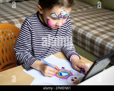 Kid drawing indoor, little girl with painted mask on face studying at home. Cute child learns to draw at table in room. Preschooler and color picture Stock Photo