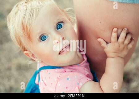 Cute blonde Caucasian girl toddler with blue eyes holding mom and looking at camera. Closeup of curious surprised baby face. Shy toddler child scared Stock Photo