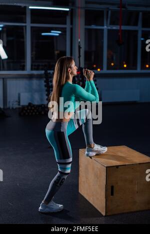 Fit young woman doing box jumping on dark background with copy space. High jump on one foot on a box. Beautiful athlete performs a circular workout Stock Photo