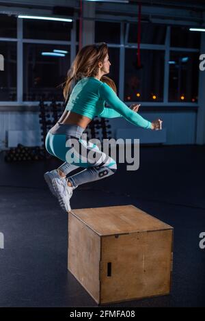 Fit young woman doing box jumping on dark background with copy space. High jump on one foot on a box. Beautiful athlete performs a circular workout Stock Photo