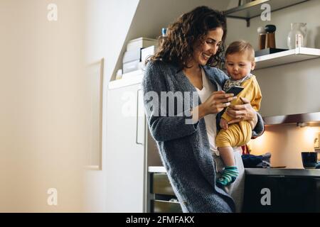 Smiling woman using mobile phone while carrying baby. Mother holding son and texting on cell phone at home. Stock Photo