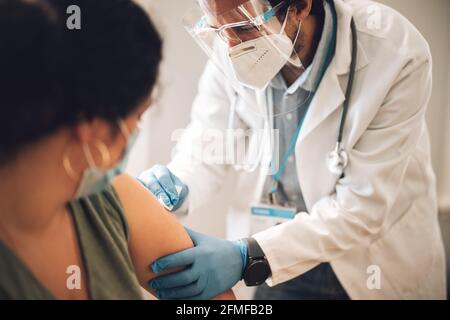 Doctor wearing face mask and shield vaccinating woman at home. Frontline workers giving covid vaccine to woman at her home. Stock Photo