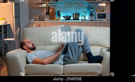 Exhausted man falling asleep while working on internet communication project using laptop computer with modern technology wireless. Caucasian male in pajamas sitting on sofa late at night in kitchen Stock Photo