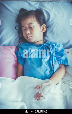 Illness asian child admitted in hospital with saline intravenous (IV) on hand. Girl sleeping at comfortable equipped hospital room with sunlight. Heal Stock Photo