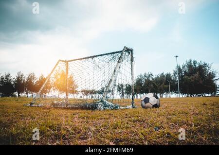 Old vacant soccer goal gate in neglected on rural grass field, football is nearby. Sports field with rusty goal and net, outdoor with bright sunlight Stock Photo