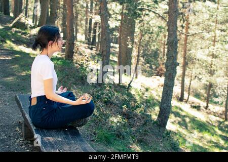 Side view of a young woman sitting on a bench while doing yoga in the forest. Stock Photo