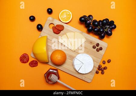 Wooden board with different cheeses, oranges, nuts, dried apricots, grapes and honey. Top view, flat lay. Orange background. Stock Photo