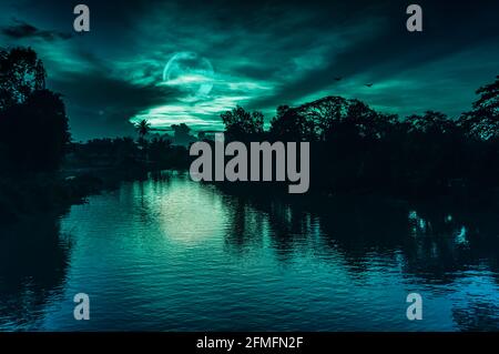 Beautiful landscape of night sky and full moon behind partial cloudy above silhouettes of trees at riverside. Serenity nature background, outdoor at n Stock Photo