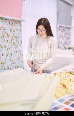 Brunette dressmaker woman in eyeglasses cutting yellow fabrics. Professional instrument concept. Tailor woman cuts linen textile cloth in atelier boutique. High quality vertical photo Stock Photo