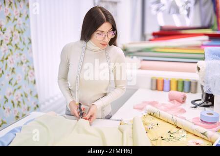 Attractive brunette seamstress woman cuts linen textile cloth in atelier boutique. Cutting yellow fabrics with blurred background. Professional instrument concept. High quality photo Stock Photo
