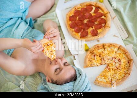 Cute girl with a towel on her head eats pizza in bed. Young woman eating pizza in bed. Life is a pleasure, body positive. Love for Italian food. Food Stock Photo