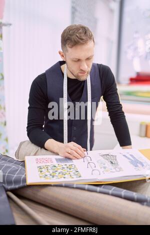 Attractive designer, craftsman or tailor in atelier. Young caucasian clothier in vest and measure tape looking through the catalogue in textile store or drapery. Garment sewing shop concept Stock Photo