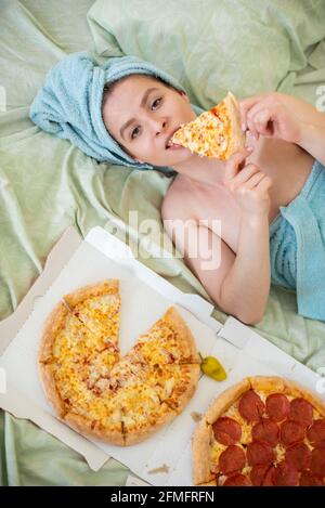 Cute girl with a towel on her head eats pizza in bed. Young woman eating pizza in bed. Life is a pleasure, body positive. Love for Italian food. Food Stock Photo