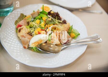 Cauliflower, greens, canned corn and fish salad with fork and spoon on served table