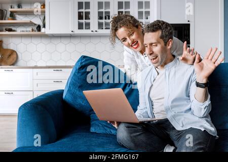 Young happy couple using laptop talking online with friends and family at home, smiling husband and wife millennial laughing and waving hello, sitting Stock Photo