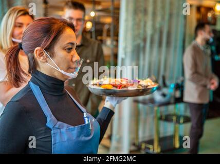 BROVARY, UKRAINE - MARCH 03, 2021: Unrecognized woman waiter wearing modern antiviral protective face mask serves a dish with food in a restaurant clo Stock Photo