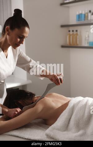 Masseur doing massage of woman back, Beautician at spa salon using gua sha metal scrapper to relieve muscle tension of female body, pressing asian tsm Stock Photo