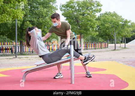 A woman doing abdominal crunches with her personal trainer at a calisthenics park wearing masks. Fitness outdoor. Stock Photo