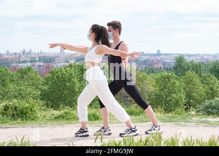 Male instructor helping young woman in a park with warrior two yoga pose wearing protective face masks. Fitness outside during pandemic. Stock Photo