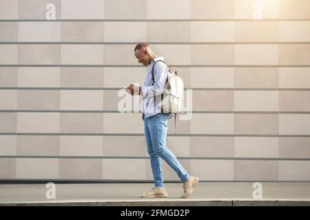 Side view of cool black guy walking along city street and using smartphone Stock Photo