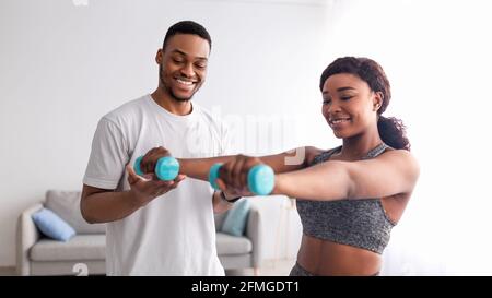 Happy black guy helping his girlfriend exercise with dumbbells at home, panorama Stock Photo